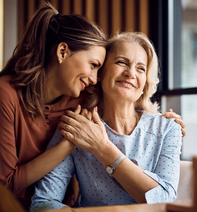 Adult daughter embracing senior mother smiling together indoors.
